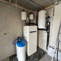 Biały piec grzewczy z zbiornikami wyrównawczymi i zmiękczaczem wody w surowym wnętrzu. Widoczne czarne rury instalacji i drabina.