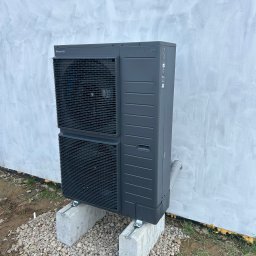 Szara pompa ciepła Panasonic Aquarea, zamontowana na betonowych blokach i żwirze przy ścianie budynku. Widoczny przewód odprowadzający skropliny.