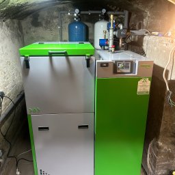 Kocioł SAS Bio Compact Plus w kotłowni z surowym, łukowym stropem. Widoczne elementy instalacji i oświetlenie robocze. Nowoczesny piec w odcieniach szarości i zieleni.