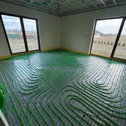 Zielone rury ogrzewania podłogowego ułożone na folii w jasnym pomieszczeniu z dwoma oknami, widok na pola w Bogdańcu. Instalacja przed wylaniem posadzki.