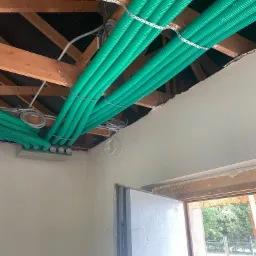 Montaż rekuperacji: widok na zielone rury wentylacyjne przymocowane do drewnianej konstrukcji pod sufitem, z widocznymi elementami instalacji i fragmentem ściany w tle.