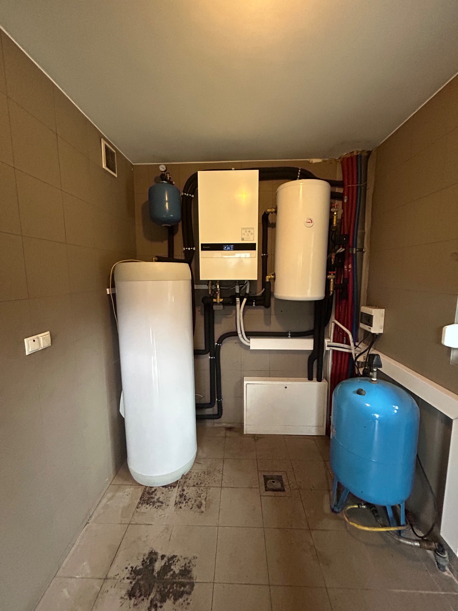 W kotłowni w Bogdańcu: biały piec gazowy, dwa białe zbiorniki, niebieski zbiornik wyrównawczy, orurowanie i instalacje na brązowych kafelkach.