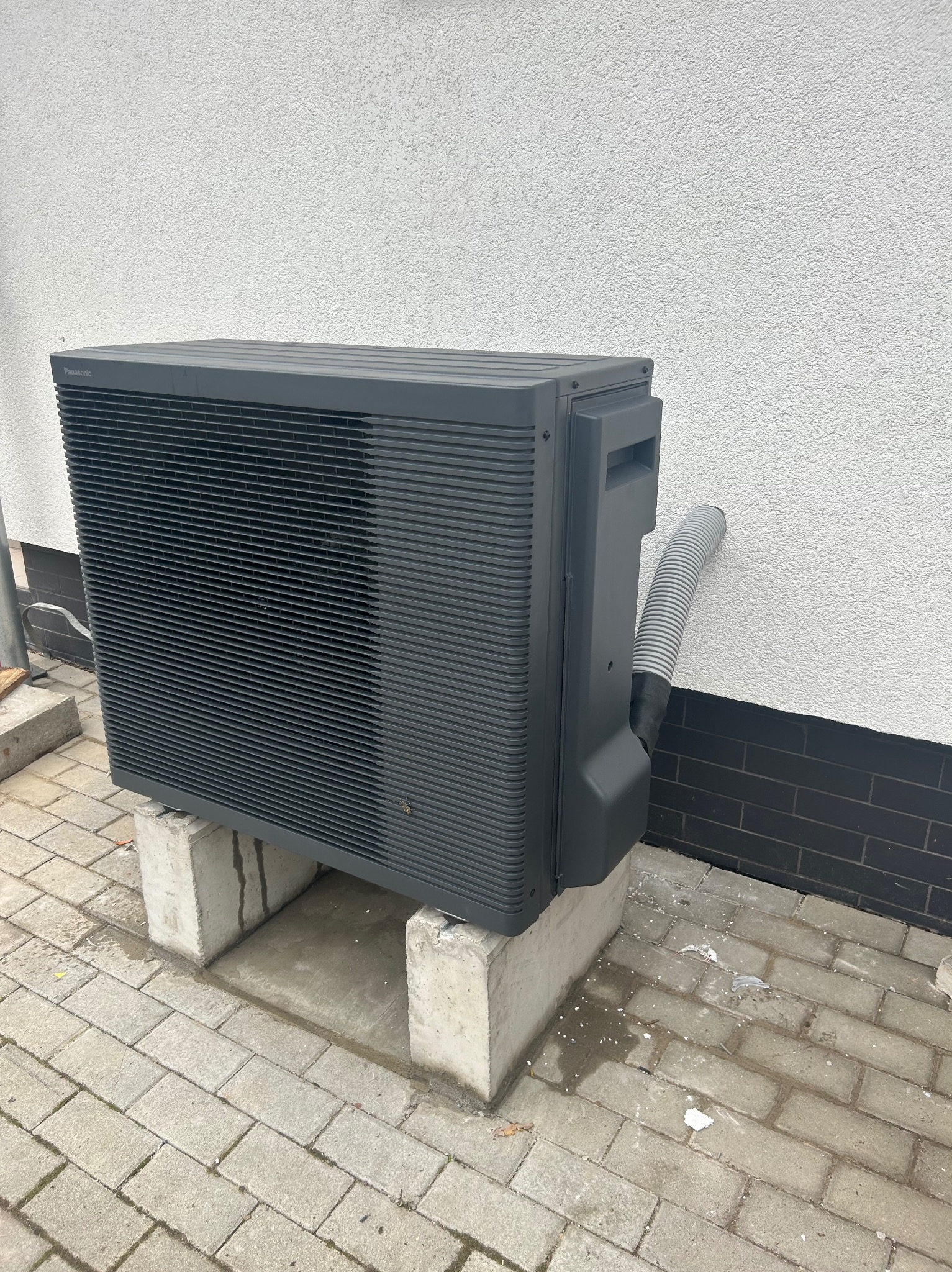 Szara jednostka zewnętrzna klimatyzacji Panasonic, stojąca na betonowych blokach przy białej ścianie budynku z widoczną szarą rurą odprowadzającą skropliny.