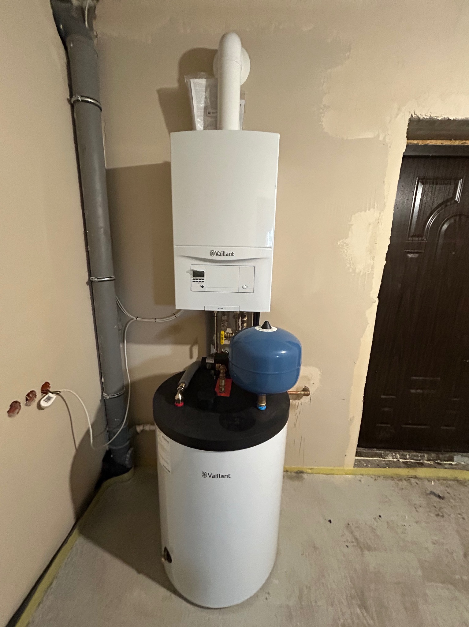 Nowo zamontowany, wiszący piec gazowy Vaillant z podłączonym zasobnikiem ciepłej wody i niebieskim naczyniem przeponowym, w surowym pomieszczeniu z widocznymi śladami po pracach instalacyjnych...