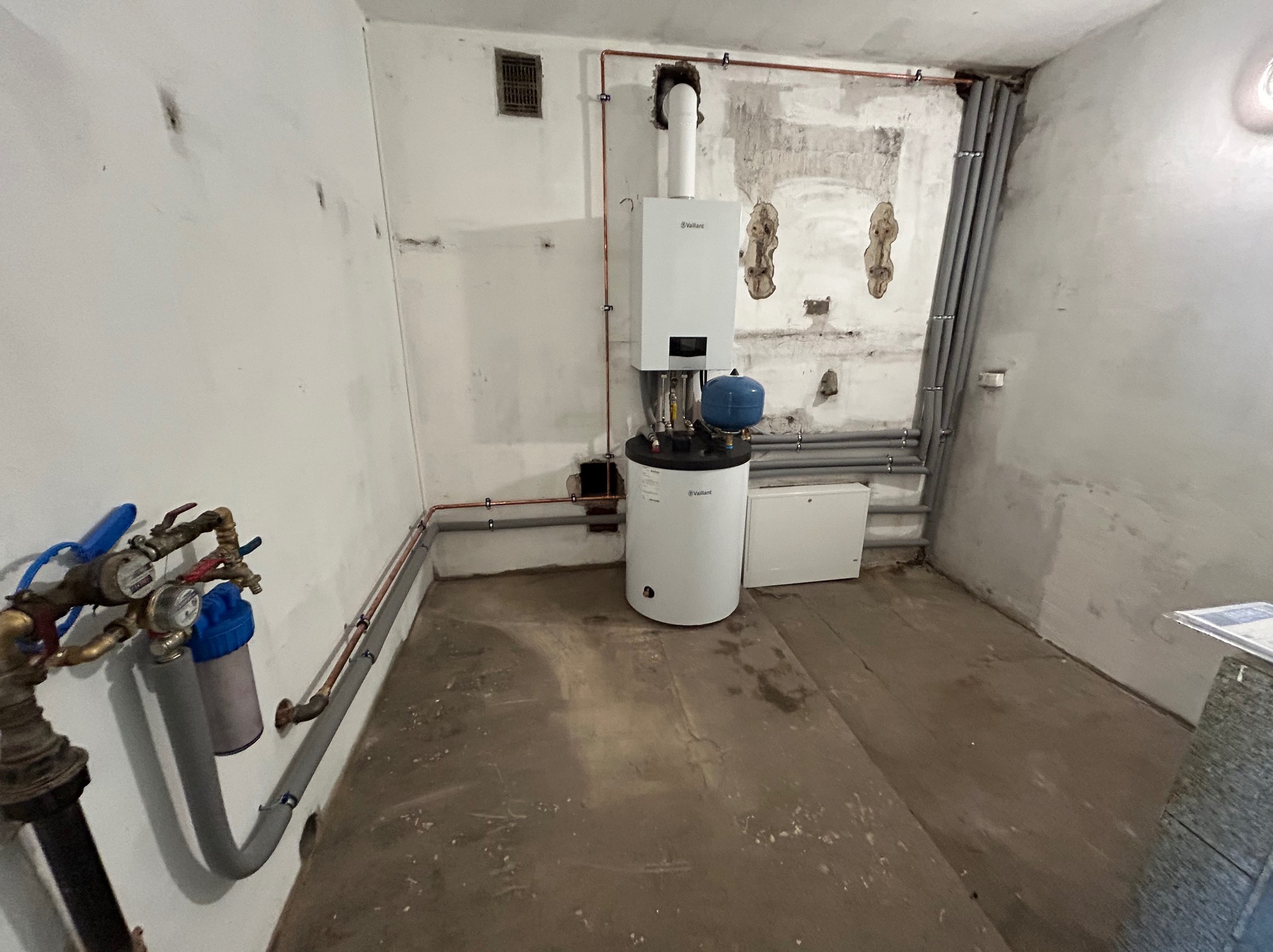Biały piec gazowy Vaillant z niebieskim zbiornikiem wyrównawczym, zamontowany w pomieszczeniu z widocznymi rurami miedzianymi i szarymi rurami PCV na ścianach o surowym wykończeniu.