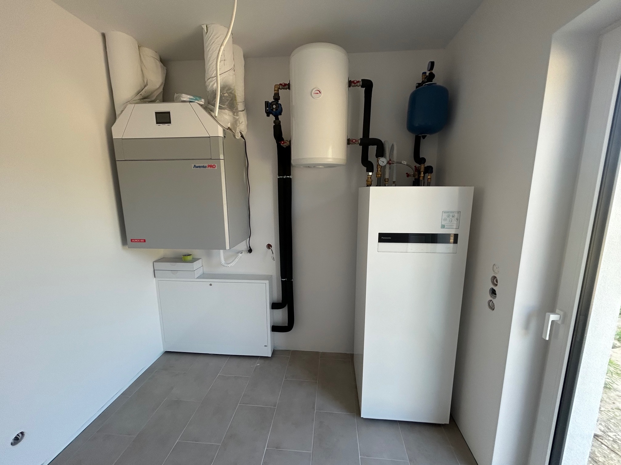 Kompaktowa kotłownia w minimalistycznym stylu: szary piec Awerenta PRO, biały zbiornik ciepłej wody, niebieskie naczynie przeponowe, czarne rury, jasne płytki podłogowe, wnęka z instalacją w nowym...
