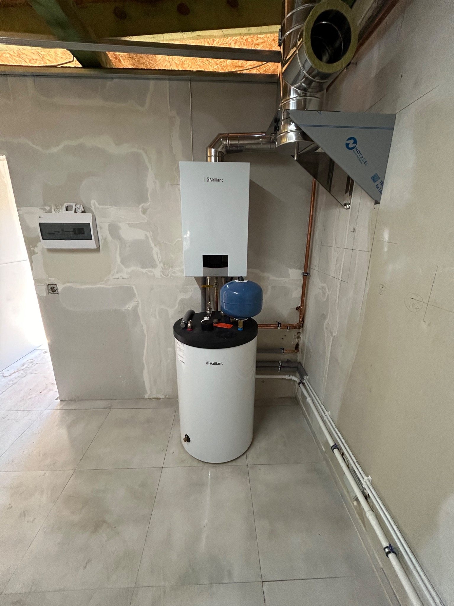 Biały piec gazowy Vaillant z niebieskim zbiornikiem wyrównawczym, rurami miedzianymi i stalowym kominem w surowym pomieszczeniu z niedokończonymi ścianami.