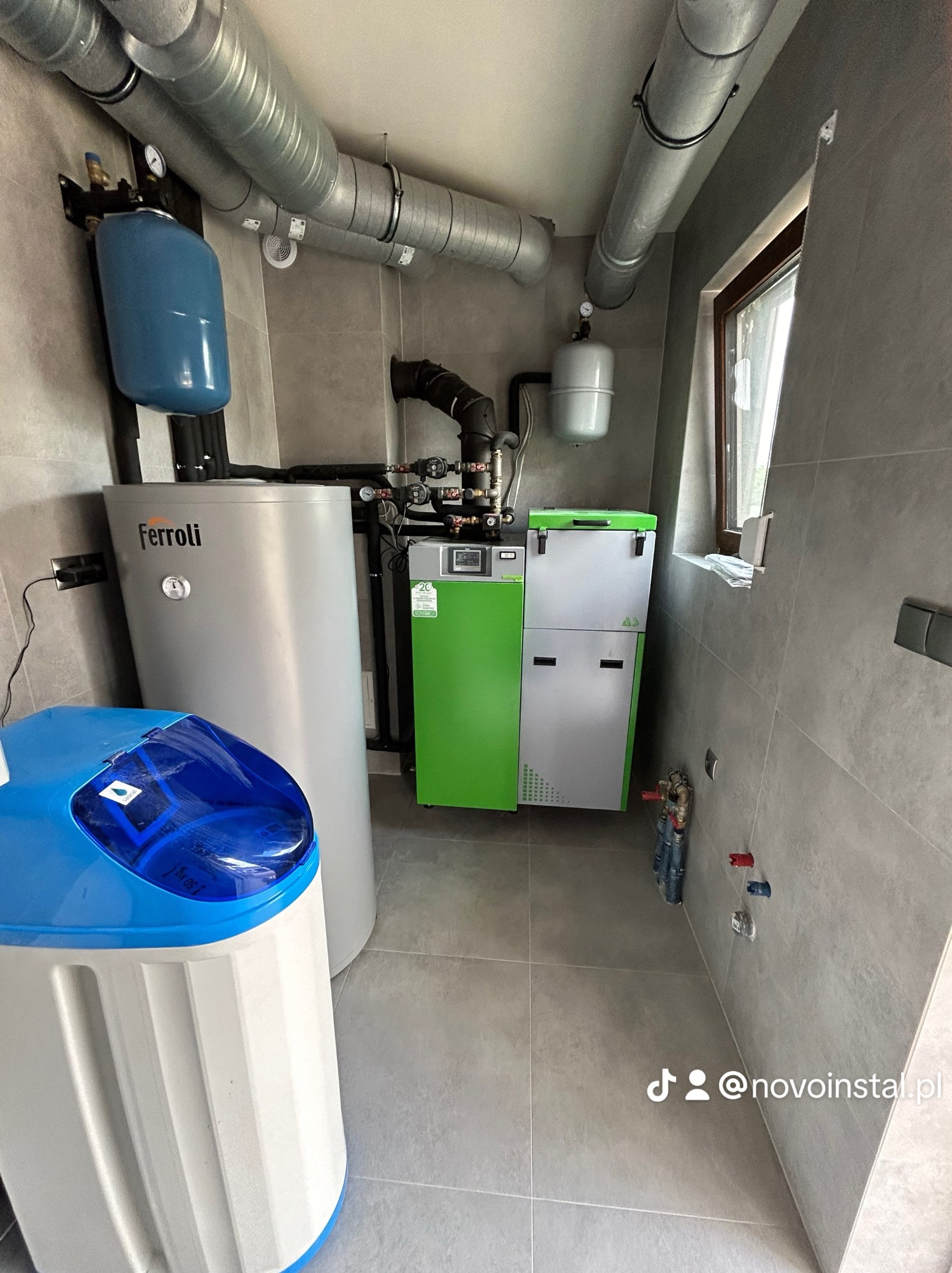 Kotłownia z piecem na pellet, zasobnikiem ciepłej wody Ferroli i zmiękczaczem wody w pomieszczeniu wykończonym szarą płytką, widoczne rury wentylacyjne pod sufitem.