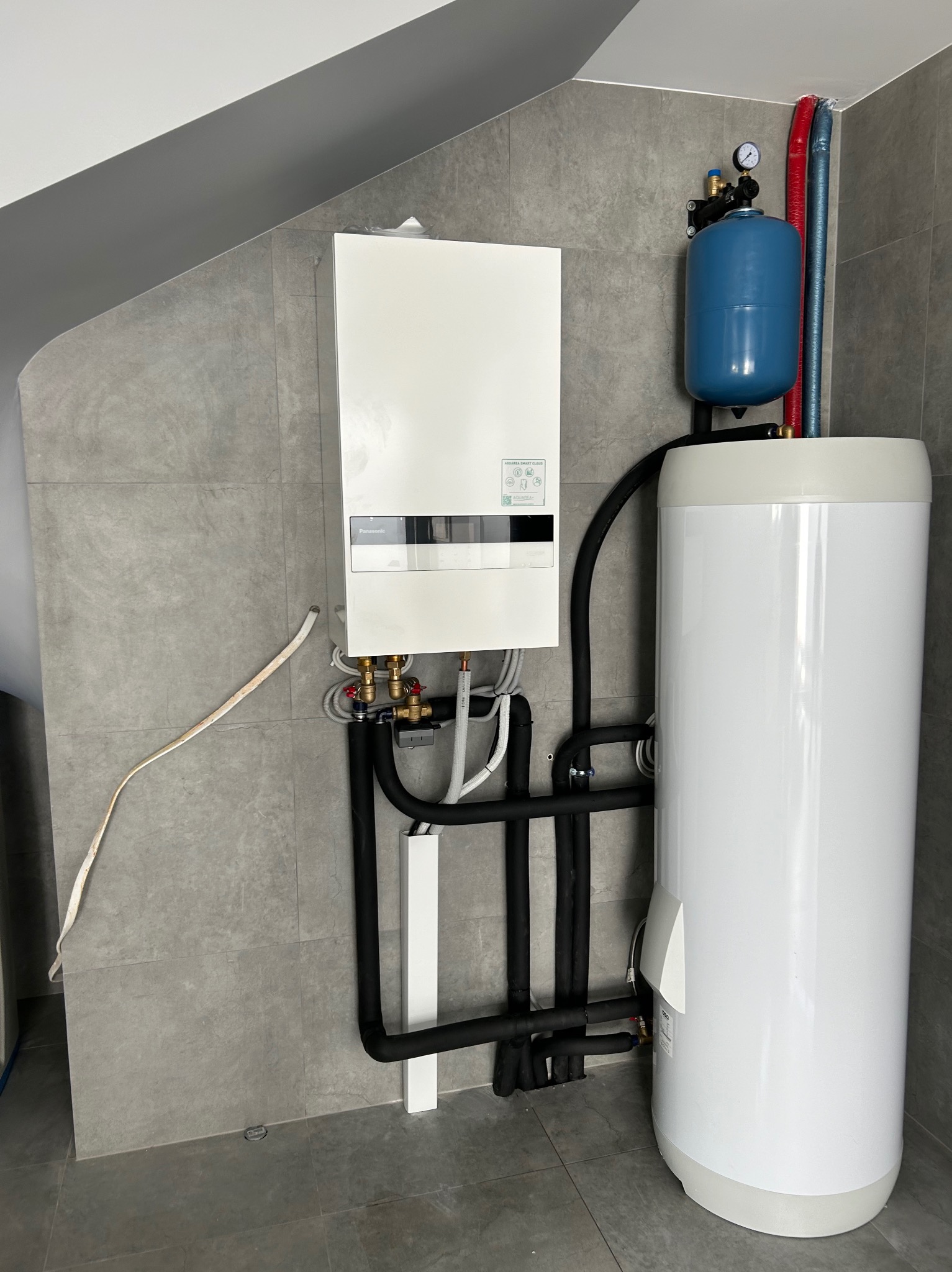 Nowoczesny, biały piec gazowy Panasonic zamontowany na szarej, kafelkowanej ścianie, z podłączonym zbiornikiem na wodę, rury w izolacji, widoczny manometr i instalacja.
