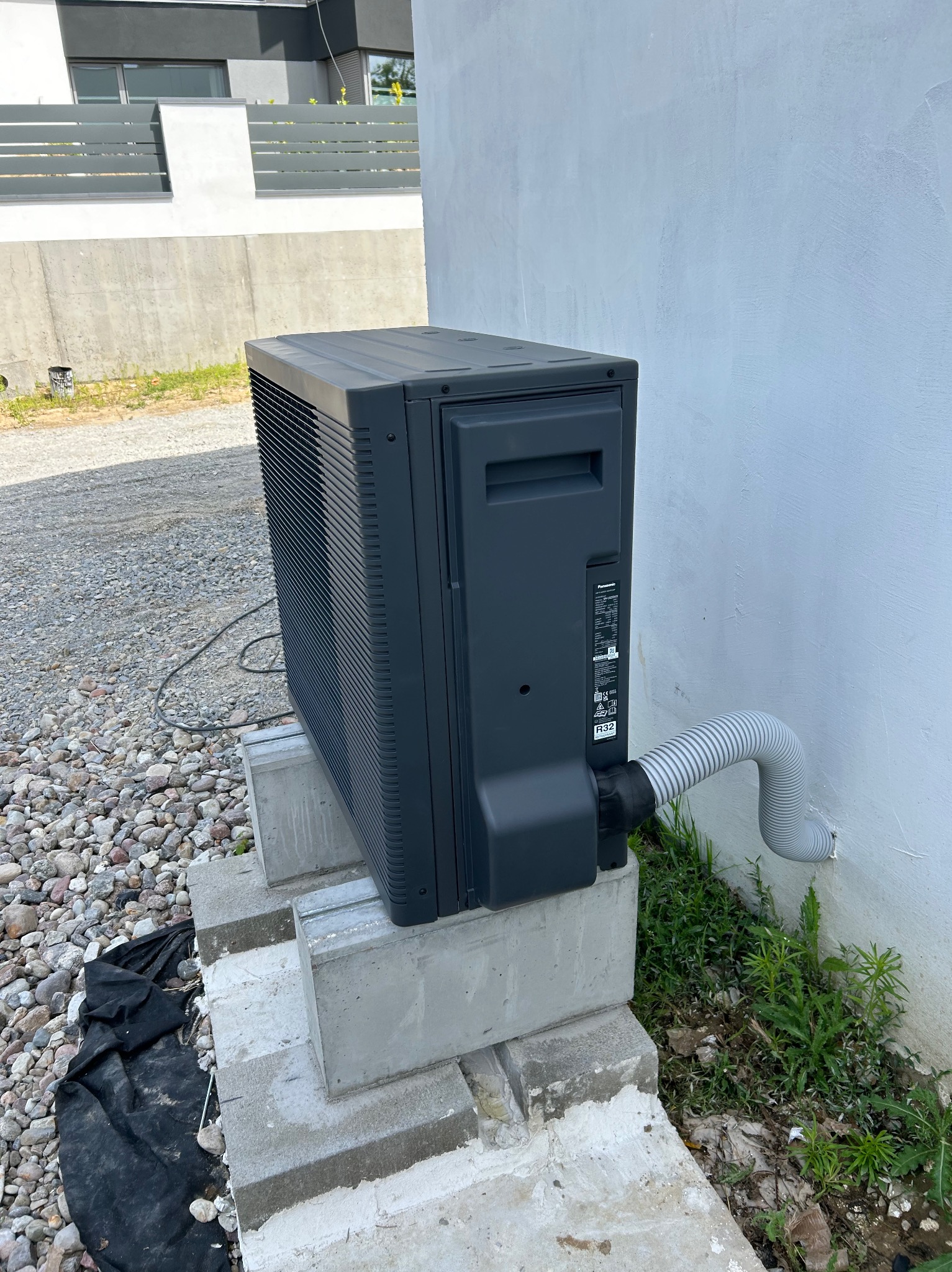 Zamontowana, szara pompa ciepła Panasonic, stojąca na betonowych bloczkach przy białej ścianie budynku, z widoczną karbowaną rurą odprowadzającą skropliny.