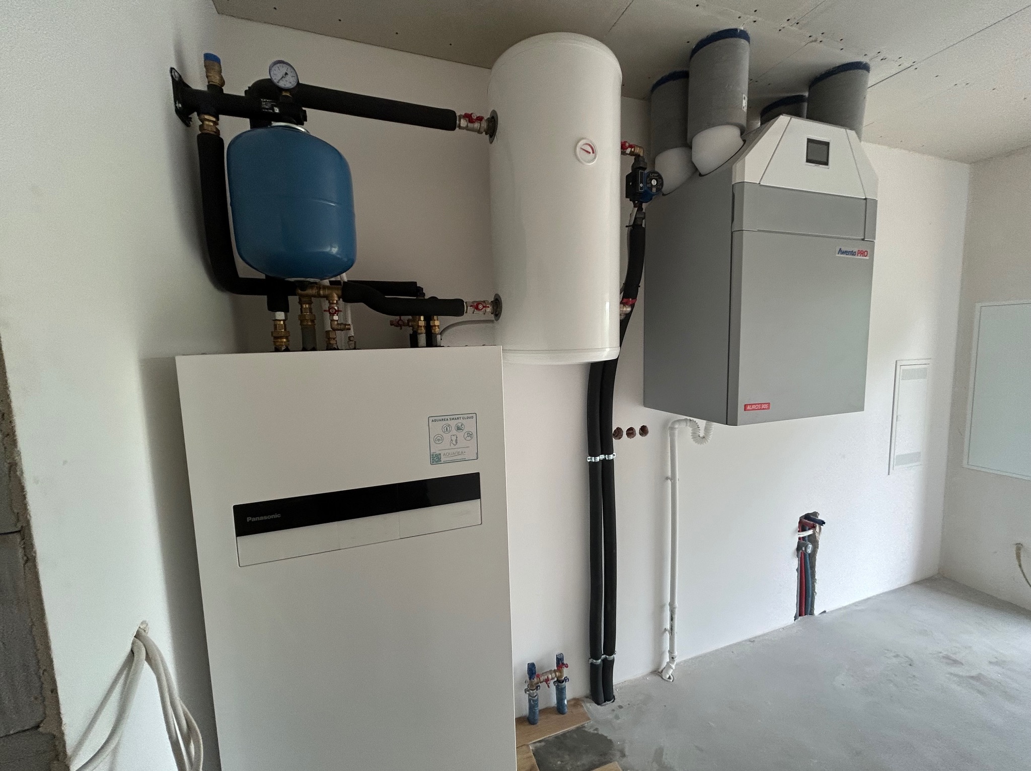Nowoczesna instalacja grzewcza w surowym wnętrzu: pompa ciepła Panasonic Aquarea, zbiornik buforowy i rekuperator Avento PRO z izolowanymi kanałami wentylacyjnymi, widoczne rury i armatura.