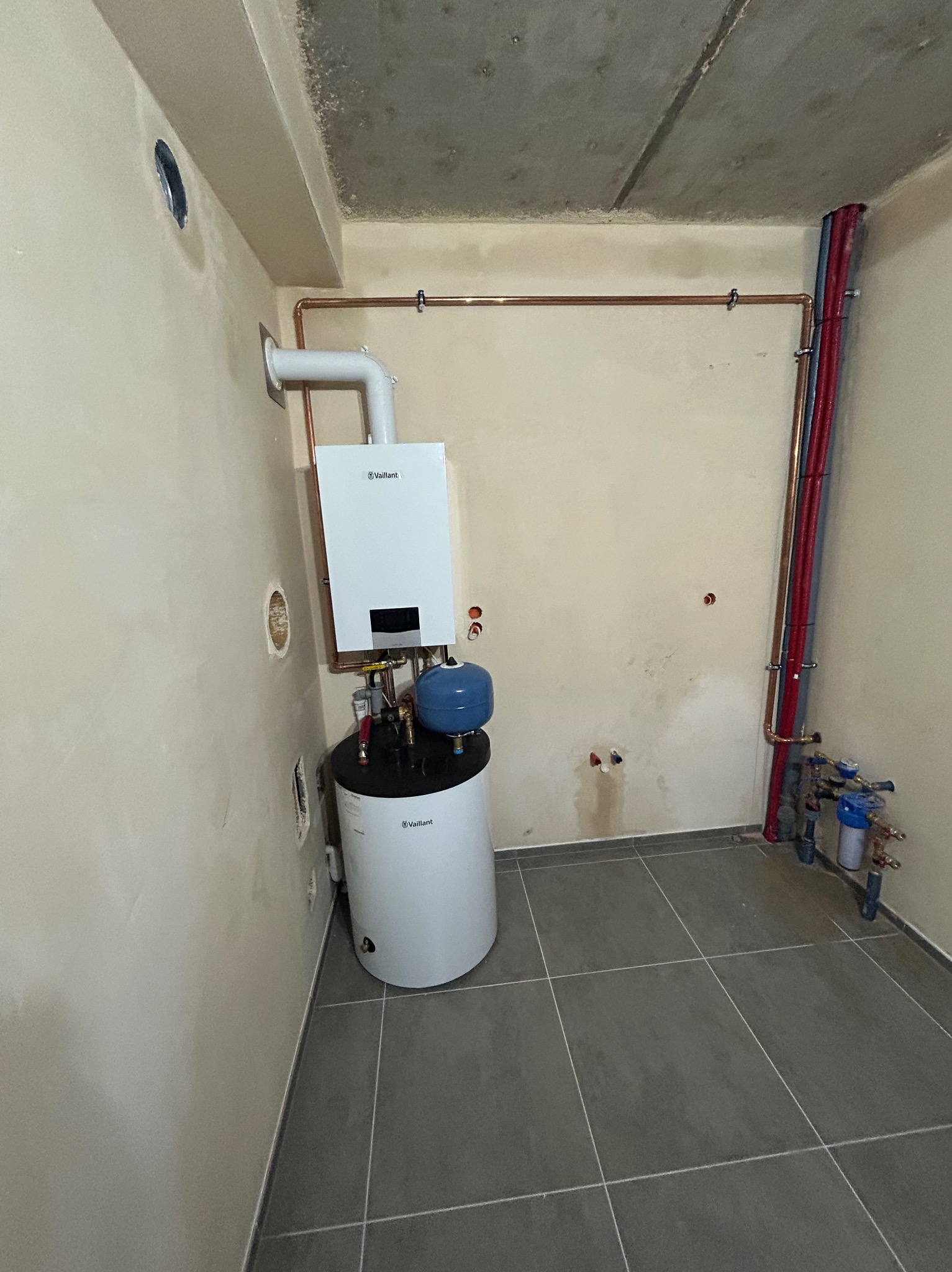 Nowy piec gazowy Vaillant z niebieskim zbiornikiem wyrównawczym zamontowany w pomieszczeniu z szarą podłogą, widoczne rury miedziane i plastikowe instalacji grzewczej.