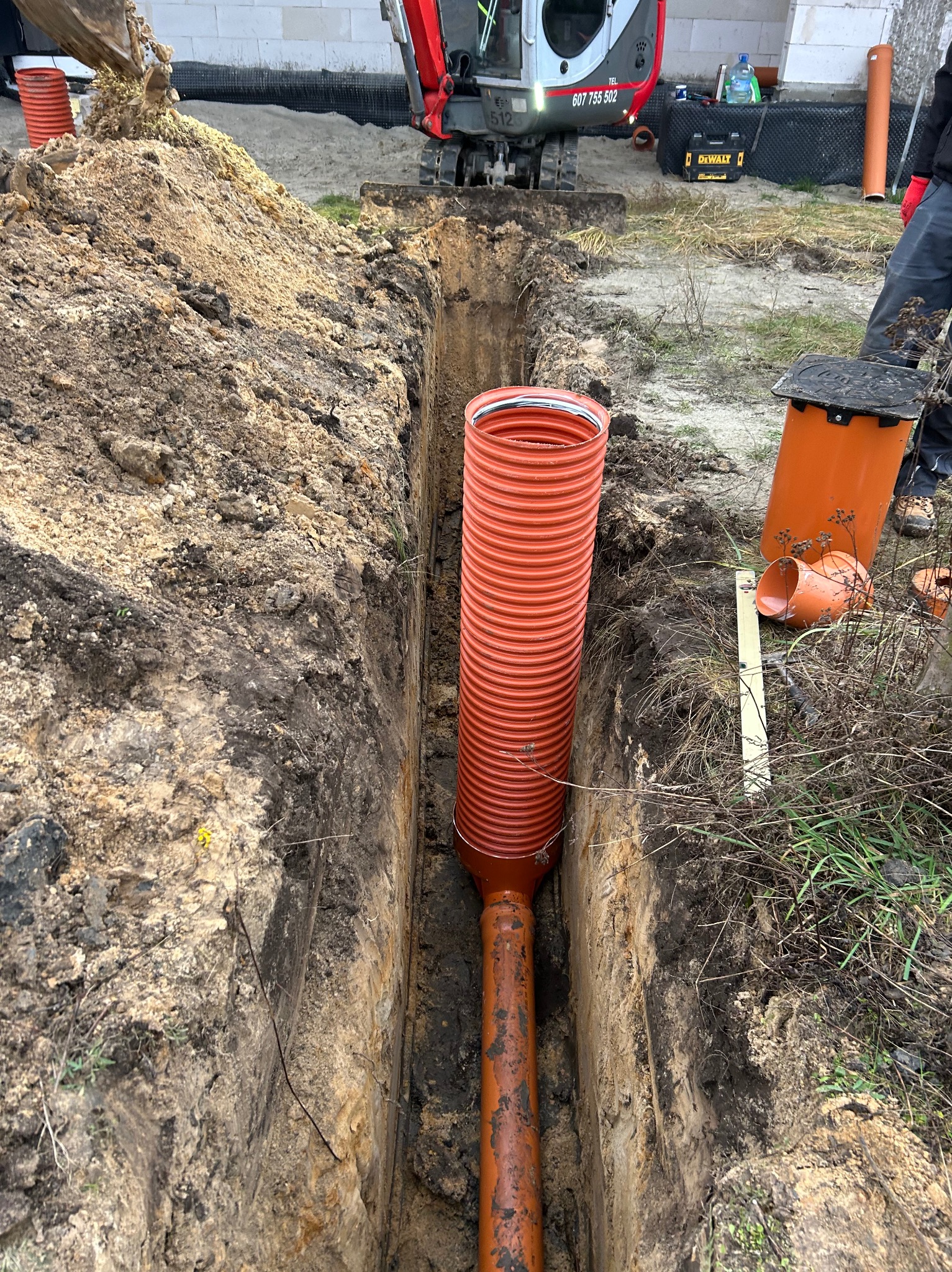 Wykop w ziemi z pomarańczową rurą karbowaną wewnątrz, widoczny mini-koparka i narzędzia DeWalt w tle, instalacja podziemna w trakcie realizacji.