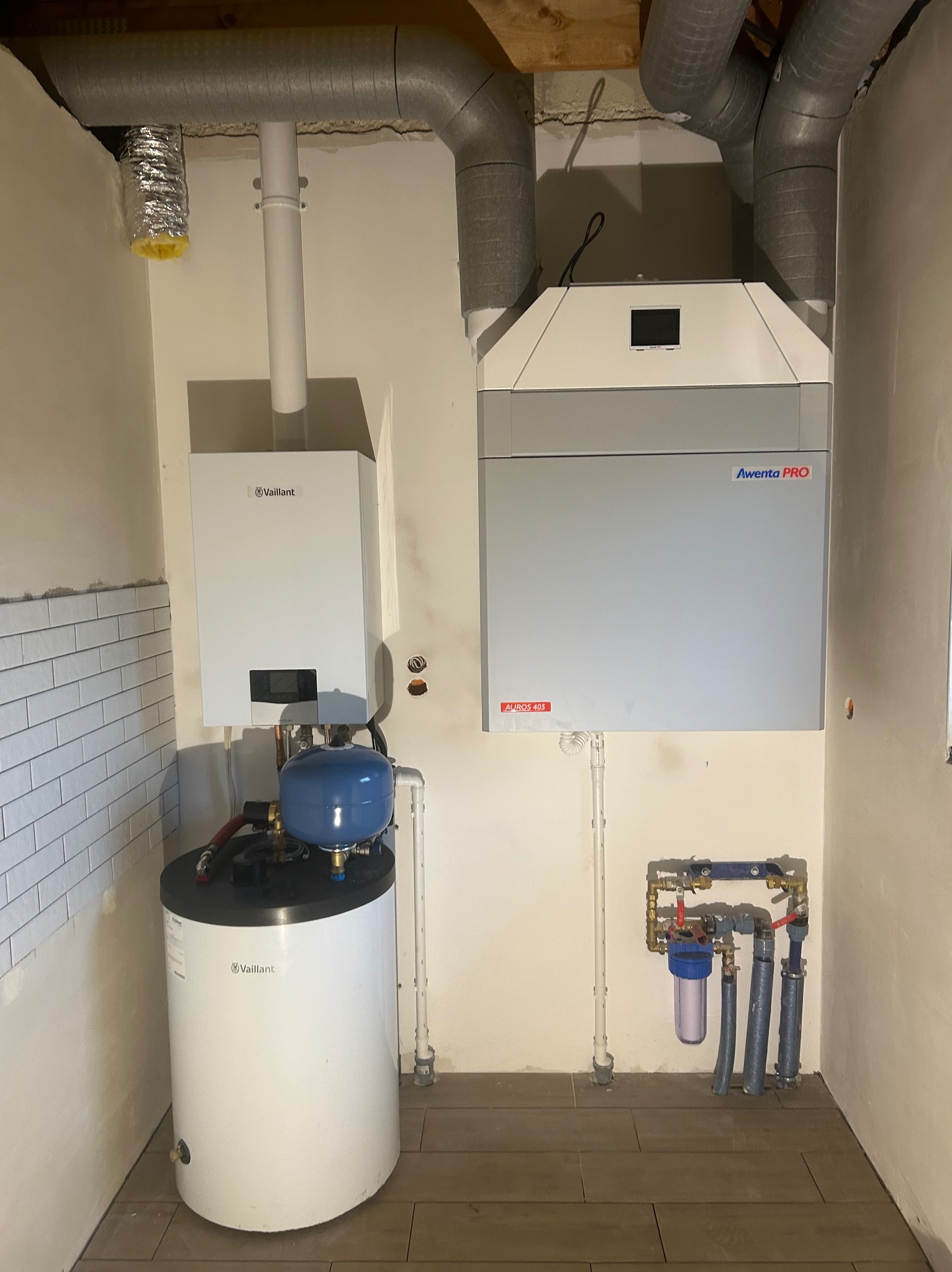 Instalacja grzewcza z piecem gazowym Vaillant, zasobnikiem ciepłej wody i rekuperatorem Aventa Pro, widoczne rury wentylacyjne i filtr wody, montaż w pomieszczeniu z wykończoną częściowo ścianą...