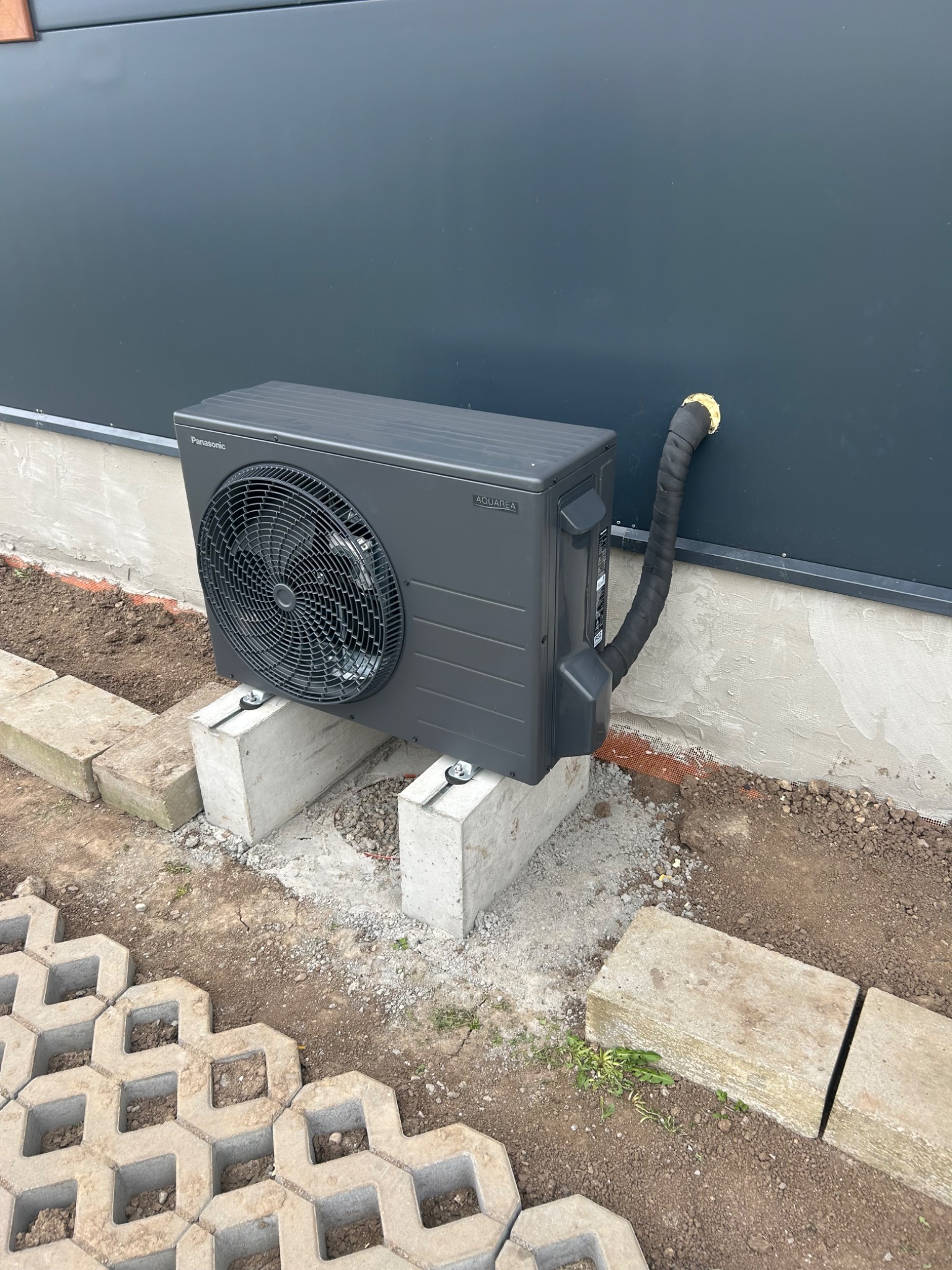 Zewnętrzna jednostka pompy ciepła Panasonic Aquarea zamontowana na betonowych bloczkach przy ciemnej ścianie budynku, z widocznym ocieplonym przewodem wychodzącym ze ściany.