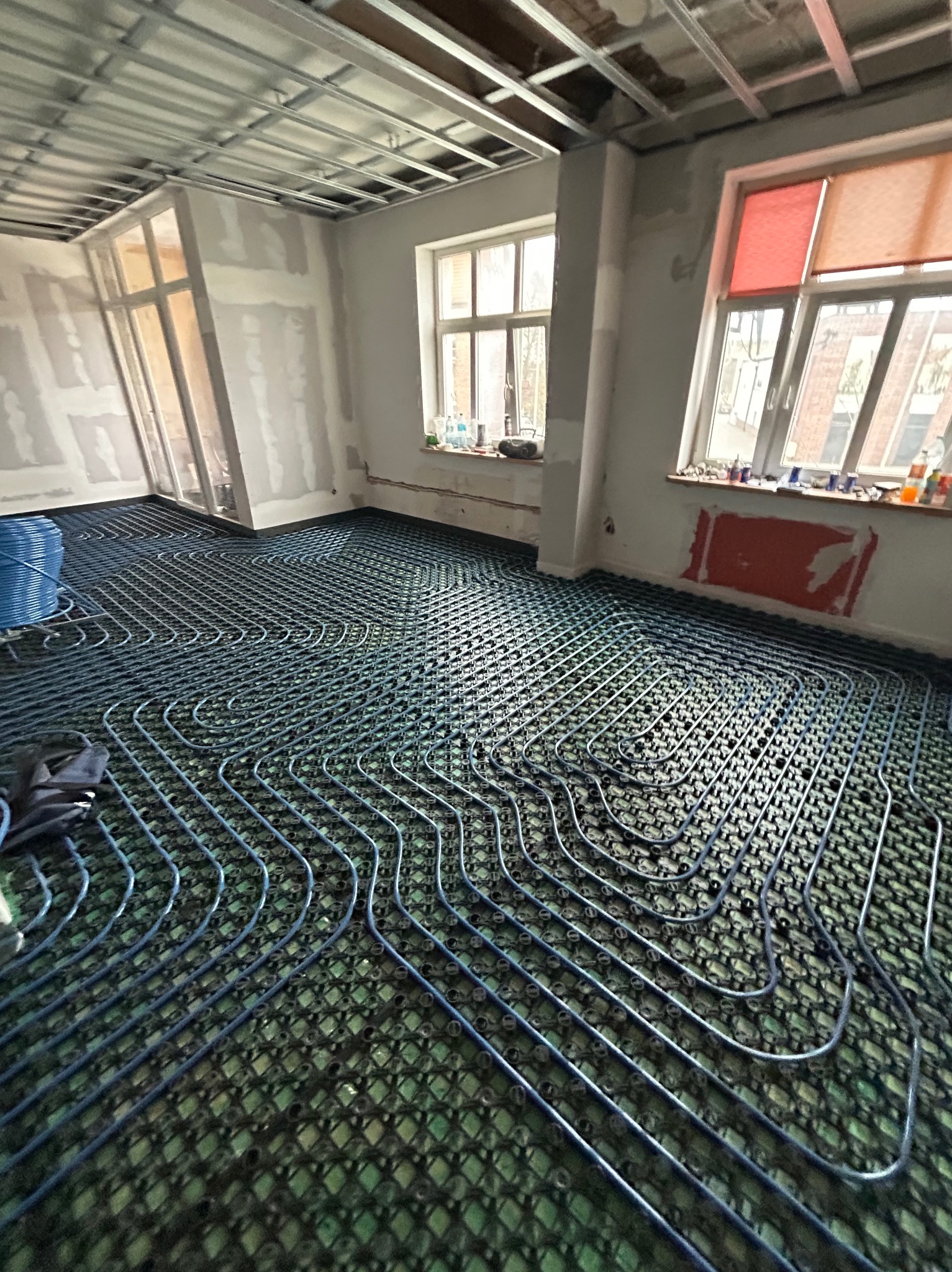 Instalacja ogrzewania podłogowego w trakcie montażu: niebieskie rurki ułożone serpentyną na zielonej macie izolacyjnej w surowym wnętrzu z widoczną konstrukcją sufitu podwieszanego i oknami.