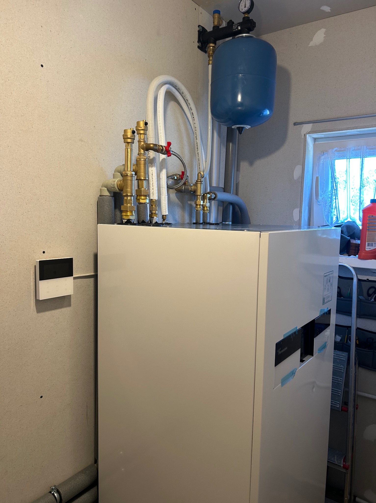 Biała pompa ciepła Panasonic z niebieskim zbiornikiem wyrównawczym, widoczne rury i zawory, na ścianie panel sterowania, w tle okno i regał z narzędziami.