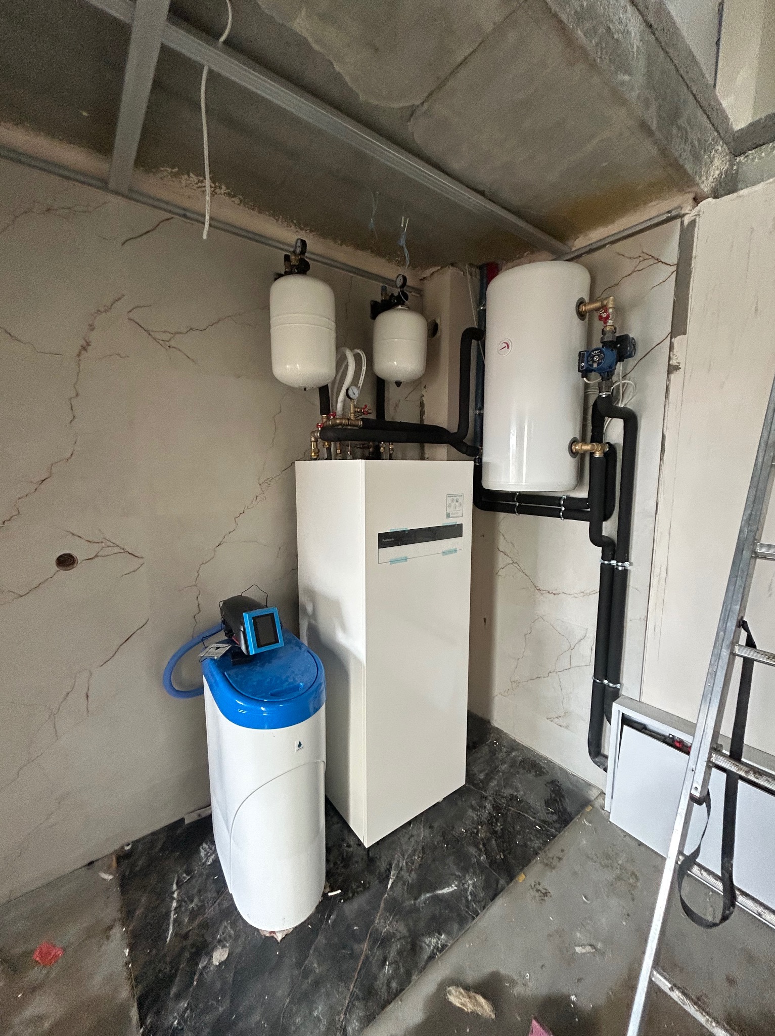 Biały piec grzewczy z zbiornikami wyrównawczymi i zmiękczaczem wody w surowym wnętrzu. Widoczne czarne rury instalacji i drabina.