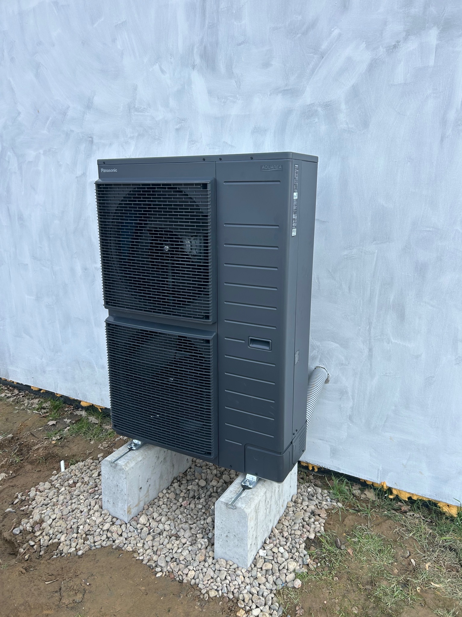 Szara pompa ciepła Panasonic Aquarea, zamontowana na betonowych blokach i żwirze przy ścianie budynku. Widoczny przewód odprowadzający skropliny.