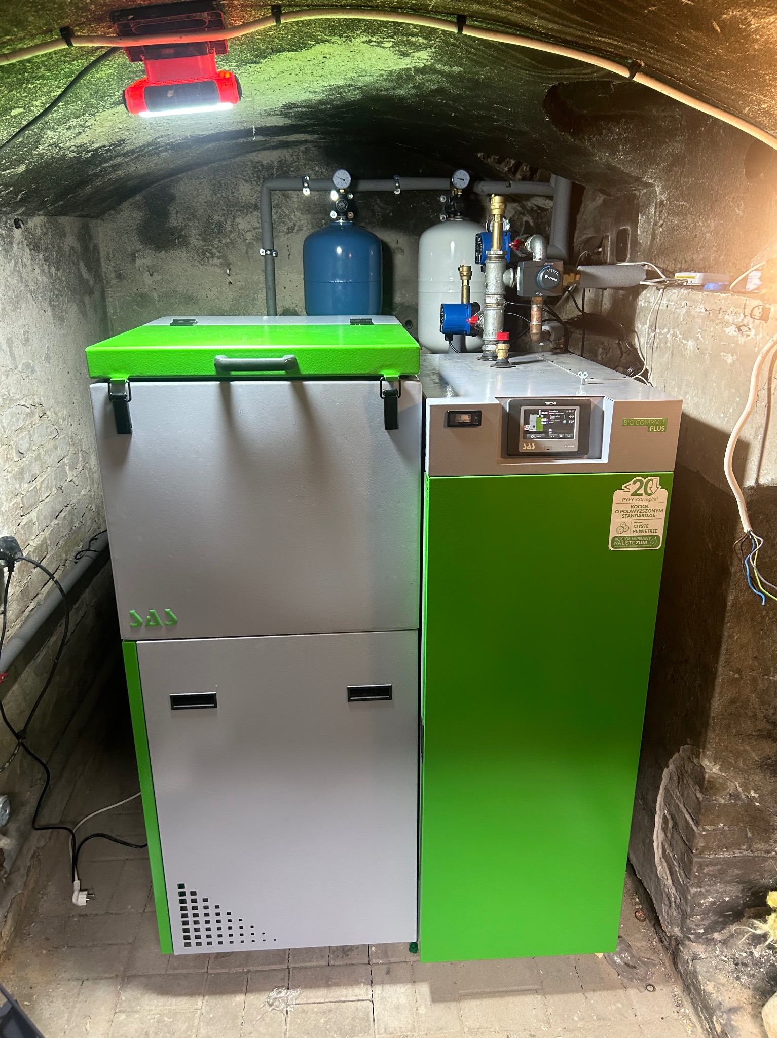 Kocioł SAS Bio Compact Plus w kotłowni z surowym, łukowym stropem. Widoczne elementy instalacji i oświetlenie robocze. Nowoczesny piec w odcieniach szarości i zieleni.