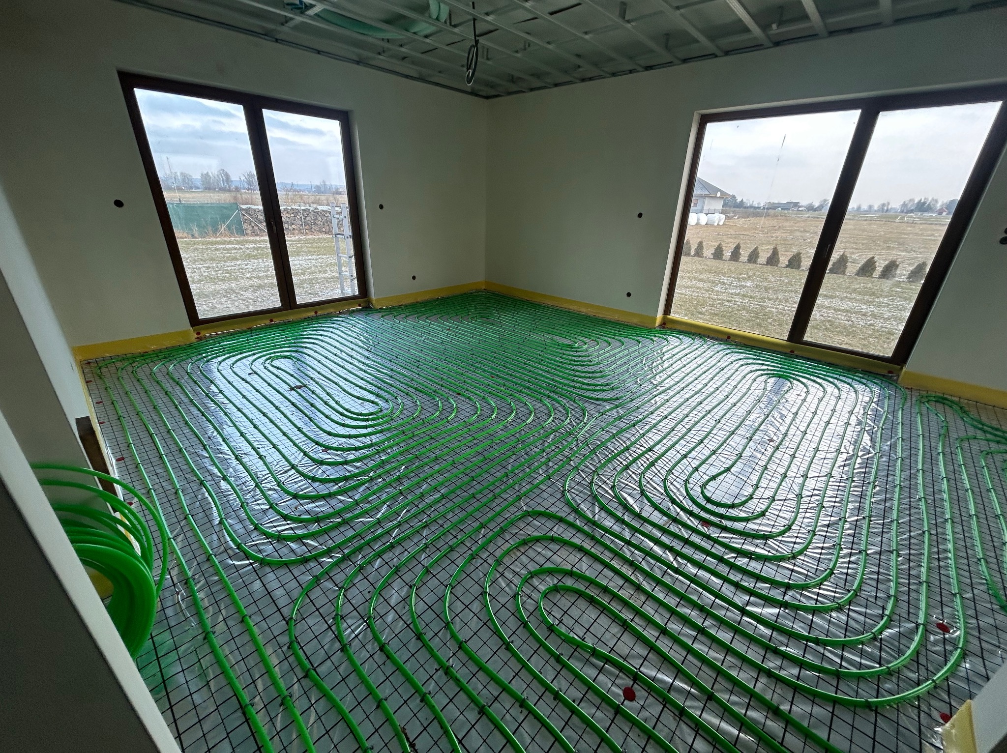 Zielone rury ogrzewania podłogowego ułożone na folii w jasnym pomieszczeniu z dwoma oknami, widok na pola w Bogdańcu. Instalacja przed wylaniem posadzki.
