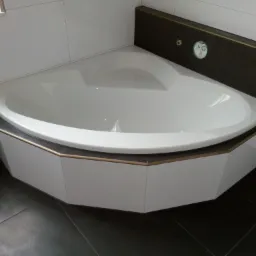 Narożna wanna z obudową w kolorze białym i brązowymi akcentami, otoczona białymi płytkami ściennymi i ciemnoszarą podłogą z płytek.