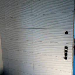 Ściana wyłożona białymi, falistymi płytkami ceramicznymi, widoczne puste otwory na gniazdka elektryczne i krzyżyki dystansowe w spoinach.