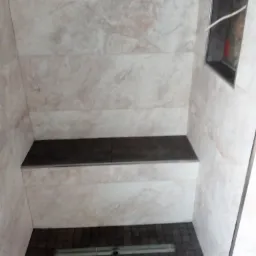 Niedokończona kabina prysznicowa z beżowymi płytkami imitującymi marmur, ciemnobrązowa mozaika podłogowa z odpływem liniowym oraz wnęka na kosmetyki w ścianie, na górze płyta kartonowo-gipsowa.