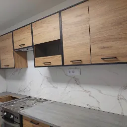 Nowoczesna kuchnia z drewnianymi szafkami z czarnymi ramami i marmurowymi płytkami ściennymi, płyta grzewcza ze stali nierdzewnej, szary blat, widoczne gniazdka elektryczne.