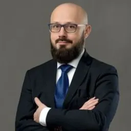 Portret mężczyzny w okularach i garniturze z niebieskim krawatem, stojącego na szarym tle z założonymi rękami.