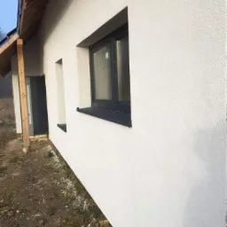 Świeżo otynkowana, biała elewacja domu jednorodzinnego z ciemnymi oknami, widoczny fragment dachu i surowy teren wokół budynku.