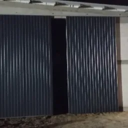 Zewnętrzne przesuwne wrota do hali magazynowej wykonane z blachy trapezowej, częściowo otwarte, oświetlone sztucznym światłem, z widocznymi paletami w tle.