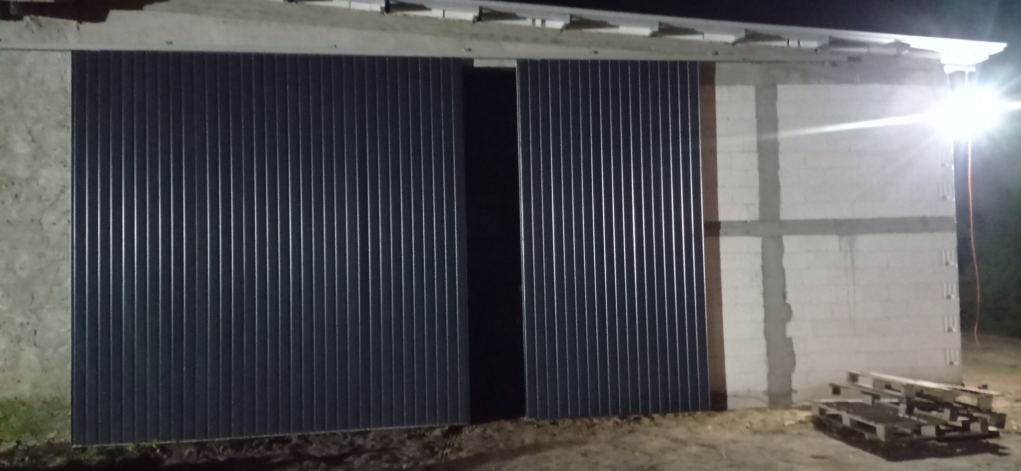 Zewnętrzne przesuwne wrota do hali magazynowej wykonane z blachy trapezowej, częściowo otwarte, oświetlone sztucznym światłem, z widocznymi paletami w tle.