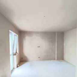 Pokój w trakcie prac wykończeniowych: ściany pokryte świeżym tynkiem, widoczne puszki elektryczne, surowa podłoga, okno z uchylonym skrzydłem, pojedynczy kabel zwisający z sufitu.