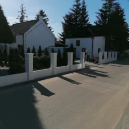 Nowoczesne białe ogrodzenie z filarami, rzucające długie cienie na asfaltową drogę; w tle białe budynki i sylwetki dwóch osób.