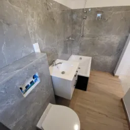 Nowoczesna łazienka w trakcie wykańczania: szare płytki imitujące marmur na ścianach, biała umywalka z szafką i czarnymi uchwytami, wanna z prysznicem, podwieszana toaleta z widocznymi instalacjami.