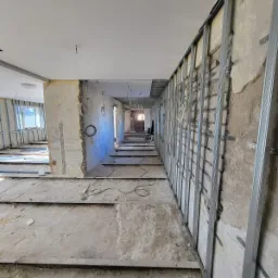 Długi korytarz w trakcie remontu, widoczne metalowe profile do montażu płyt kartonowo-gipsowych na ścianach, odsłonięta instalacja elektryczna, surowy beton na podłodze i ścianach, częściowo...