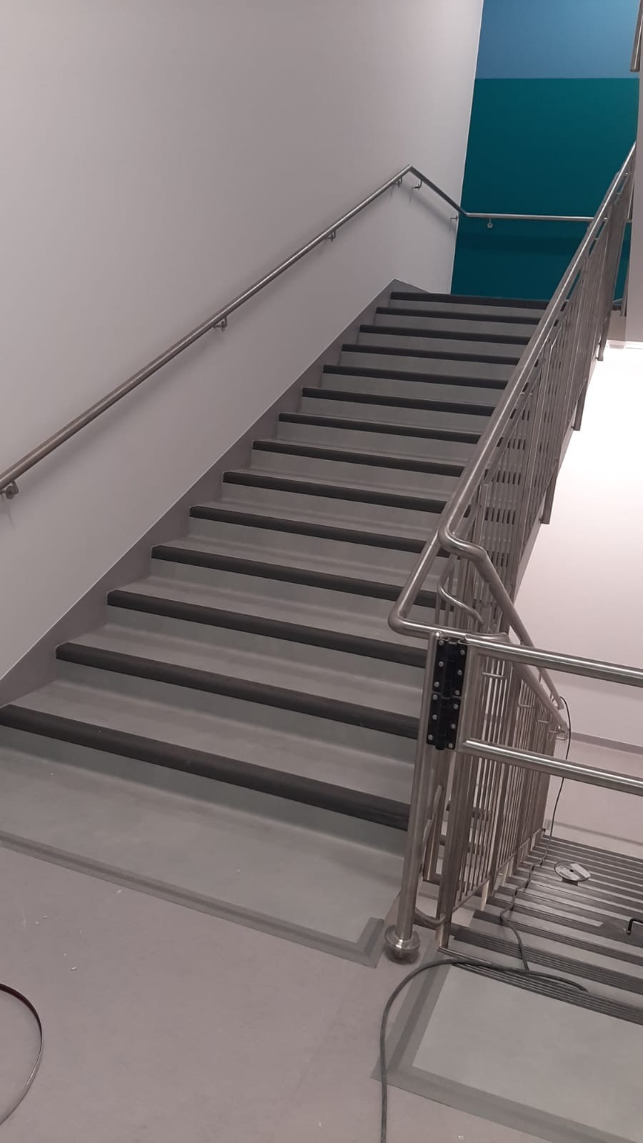 Schody wyłożone szarą wykładziną z ciemnymi pasami antypoślizgowymi, metalowe barierki i poręcze, widoczne kable na podłodze wskazujące na prace wykończeniowe.