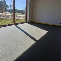 Świeżo wylana, gładka posadzka w jasnym pomieszczeniu z dużymi oknami, widoczne refleksy światła słonecznego i zabezpieczone listwy przypodłogowe.