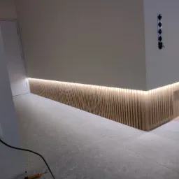 Nowoczesne wykończenie wnętrza z podświetloną listwą przypodłogową z drewnianych lameli i widocznym testerem napięcia na podłodze.