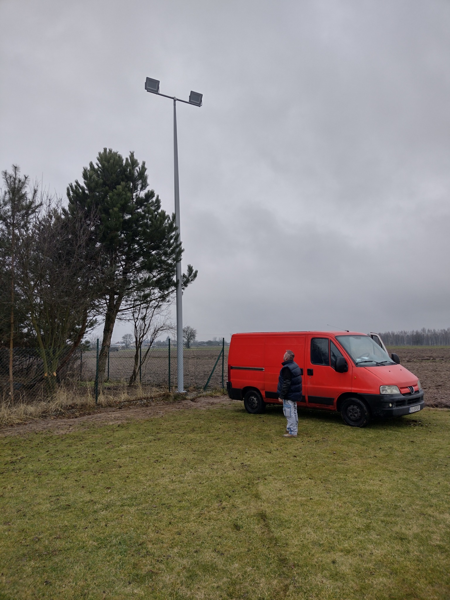 Nowo zainstalowany słup oświetleniowy z dwoma reflektorami LED na tle pochmurnego nieba, obok stoi pomarańczowy samochód dostawczy i mężczyzna w kurtce, teren trawiasty z widocznym polem w tle.