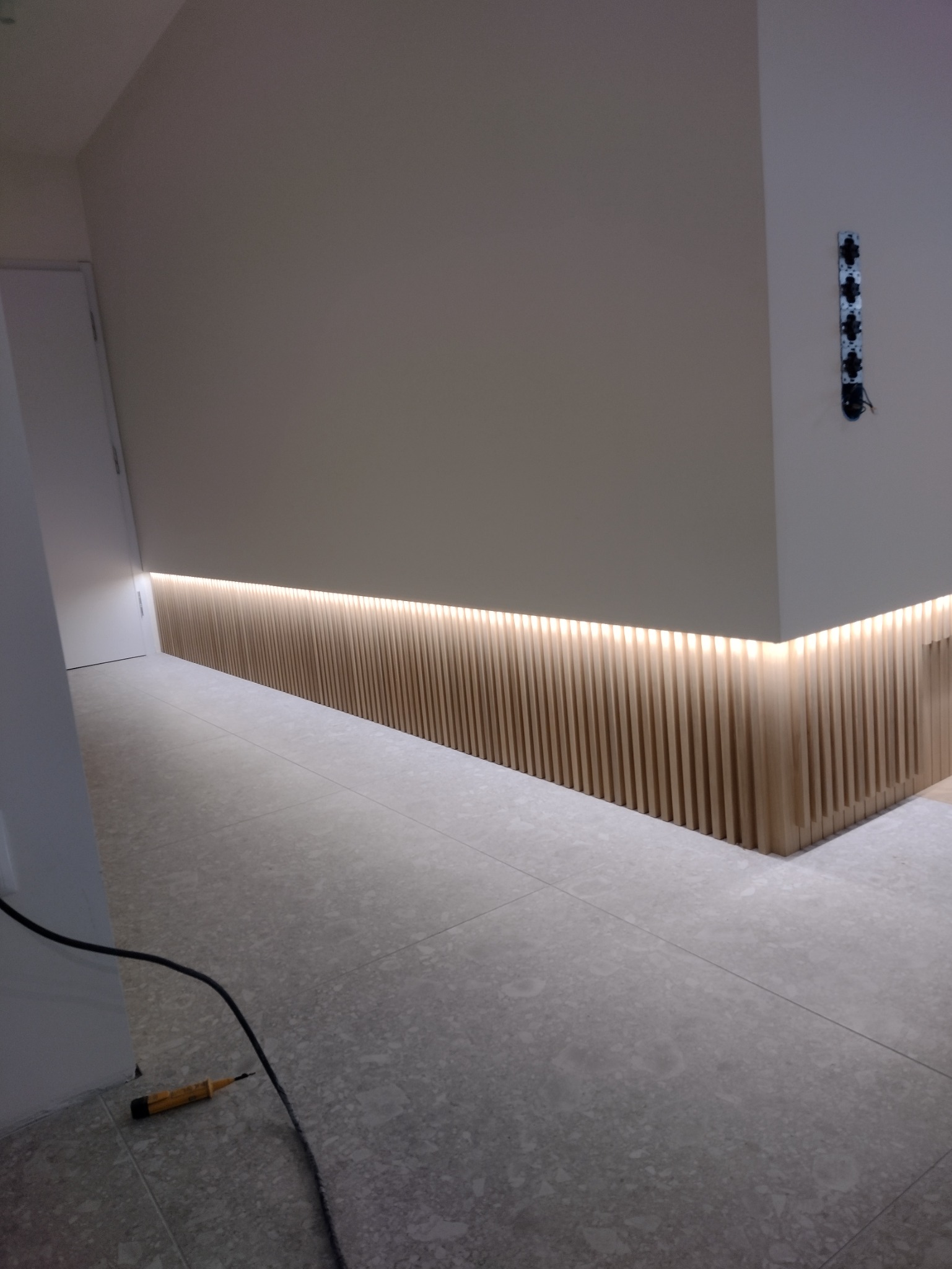 Nowoczesne wykończenie wnętrza z podświetloną listwą przypodłogową z drewnianych lameli i widocznym testerem napięcia na podłodze.