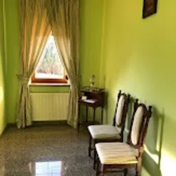 Jasnozielony pokój z dwoma krzesłami z kremowymi siedziskami, stolikiem z lampką i oknem z beżowymi zasłonami w trójkątnym układzie, marmurowa podłoga.