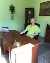 Kobieta w zielonej sukience siedzi przy biurku w gabinecie z zielonymi ścianami, na biurku leżą książki i srebrna papierośnica, w tle ciemna szafka i obraz.