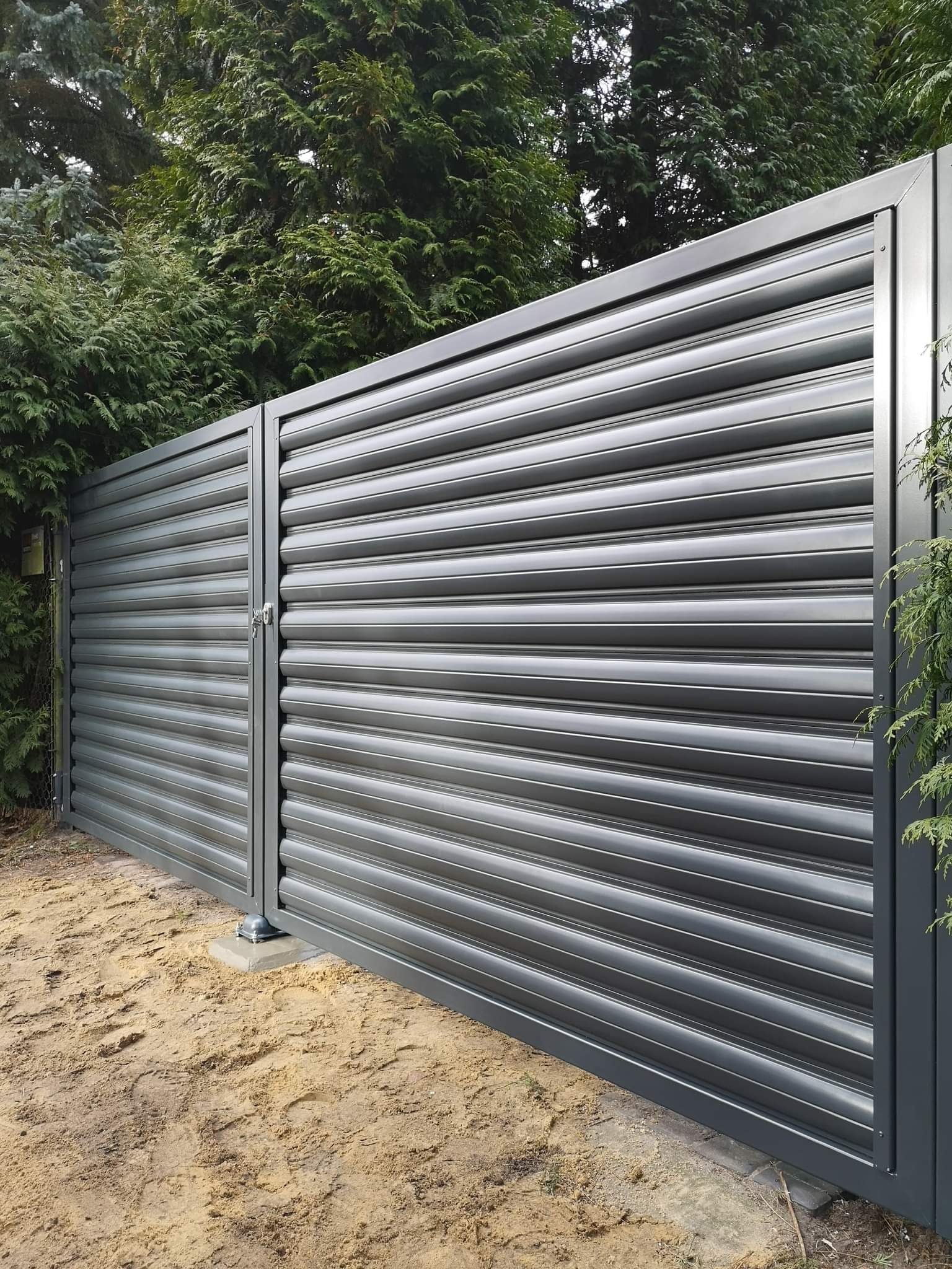 Nowoczesna, pozioma brama przesuwna w kolorze grafitowym, z widocznym zamkiem, na tle zieleni drzew.