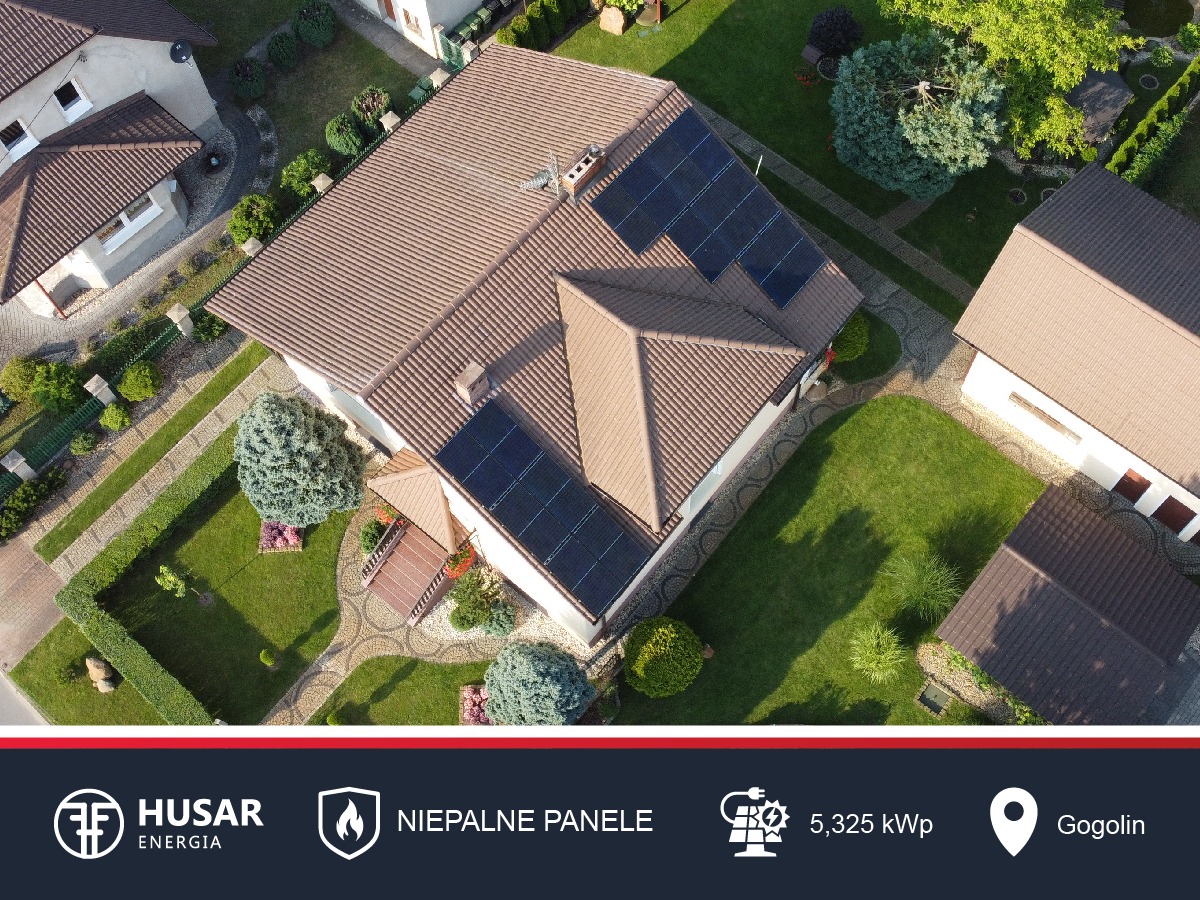 Widok z lotu ptaka na dom jednorodzinny z brązowym dachem, na którym zamontowane są panele słoneczne. Ogród z zielonym trawnikiem i ozdobnymi krzewami.