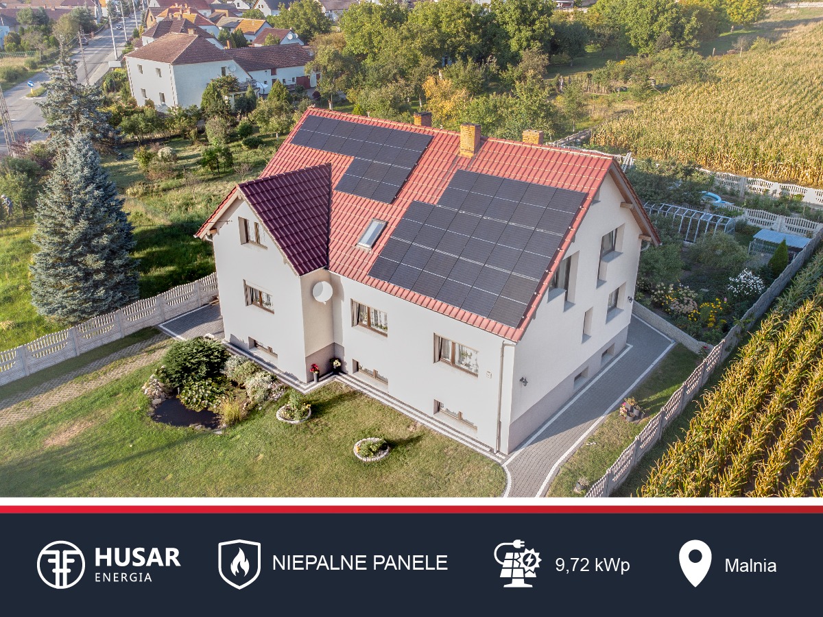 Dom jednorodzinny z czerwonym dachem i panelami słonecznymi na dachu, widok z lotu ptaka, w tle zabudowa wiejska i pole uprawne.