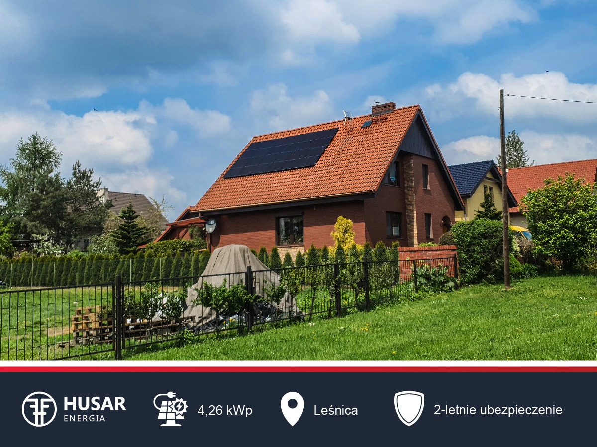 Dom jednorodzinny z czerwoną dachówką i zainstalowanymi panelami słonecznymi na dachu, widok z ogrodu z metalowym ogrodzeniem.