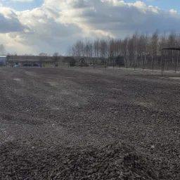 Ukończone utwardzanie terenu żwirem, widoczny fragment ogrodzenia panelowego z podmurówką, blaszany budynek gospodarczy w tle, pochmurne niebo.