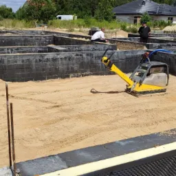 Wykop pod fundamenty z izolacją przeciwwilgociową, częściowo zasypany piaskiem, ubijany zagęszczarką. Widoczne zbrojenia, niebieski wąż i pracownicy w tle.